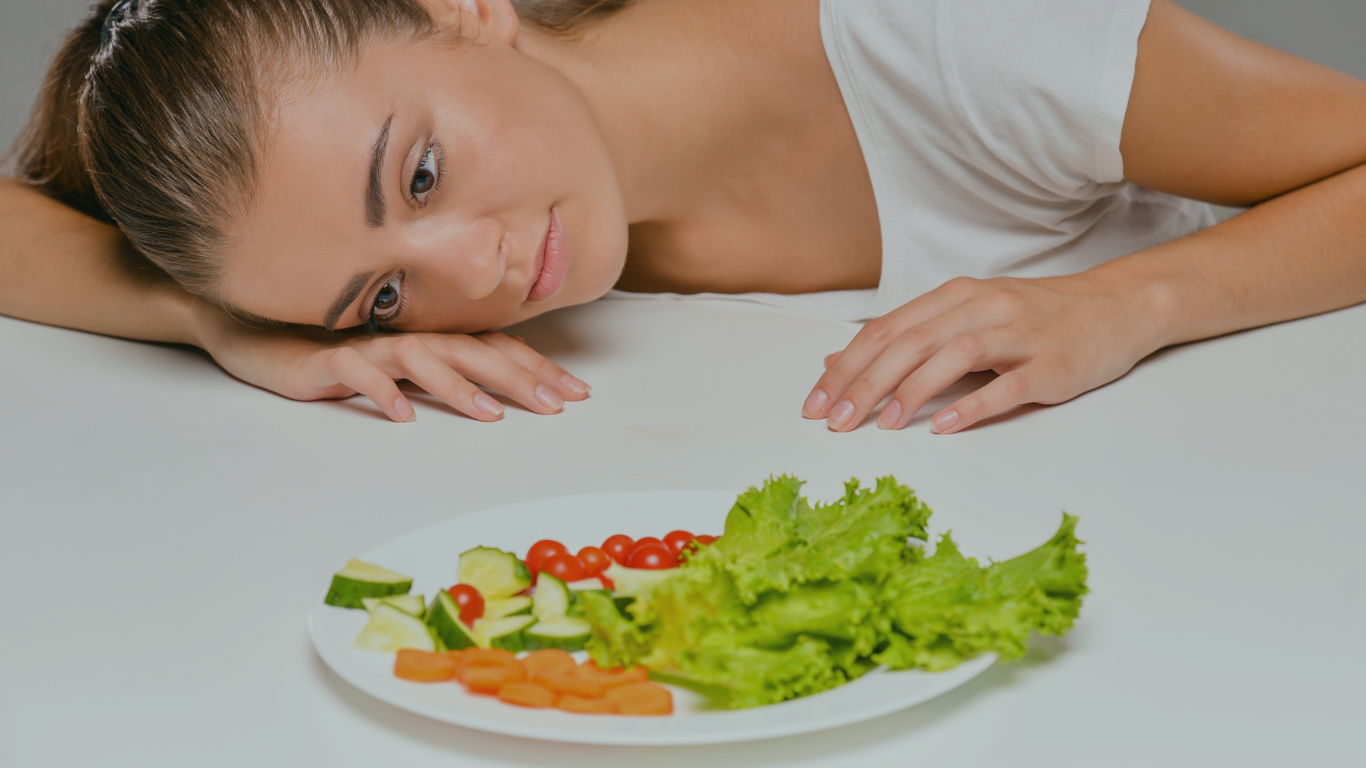 Mulher jovem deitada na mesa diante de um prato de comida, insatisfeita com suas dietas radicais